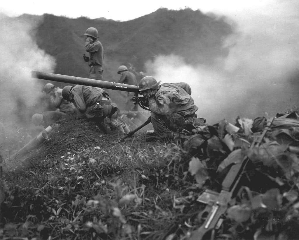 Black and white photo of U.S. Army infantry in 1951 in combat during the Korean War. The image shows soldiers from an infantry regiment firing a M20 75mm recoilless rifle from a hilltop. The smoke obscured hilltop scene is during the summer months reflecting the intensity of combat. Black and white photo of U.S. Army infantry in 1951 in combat during the Korean War. The image shows soldiers from an infantry regiment firing a M20 75mm recoilless rifle from a hilltop. The smoke obscured hilltop scene is during the summer months reflecting the intensity of combat.