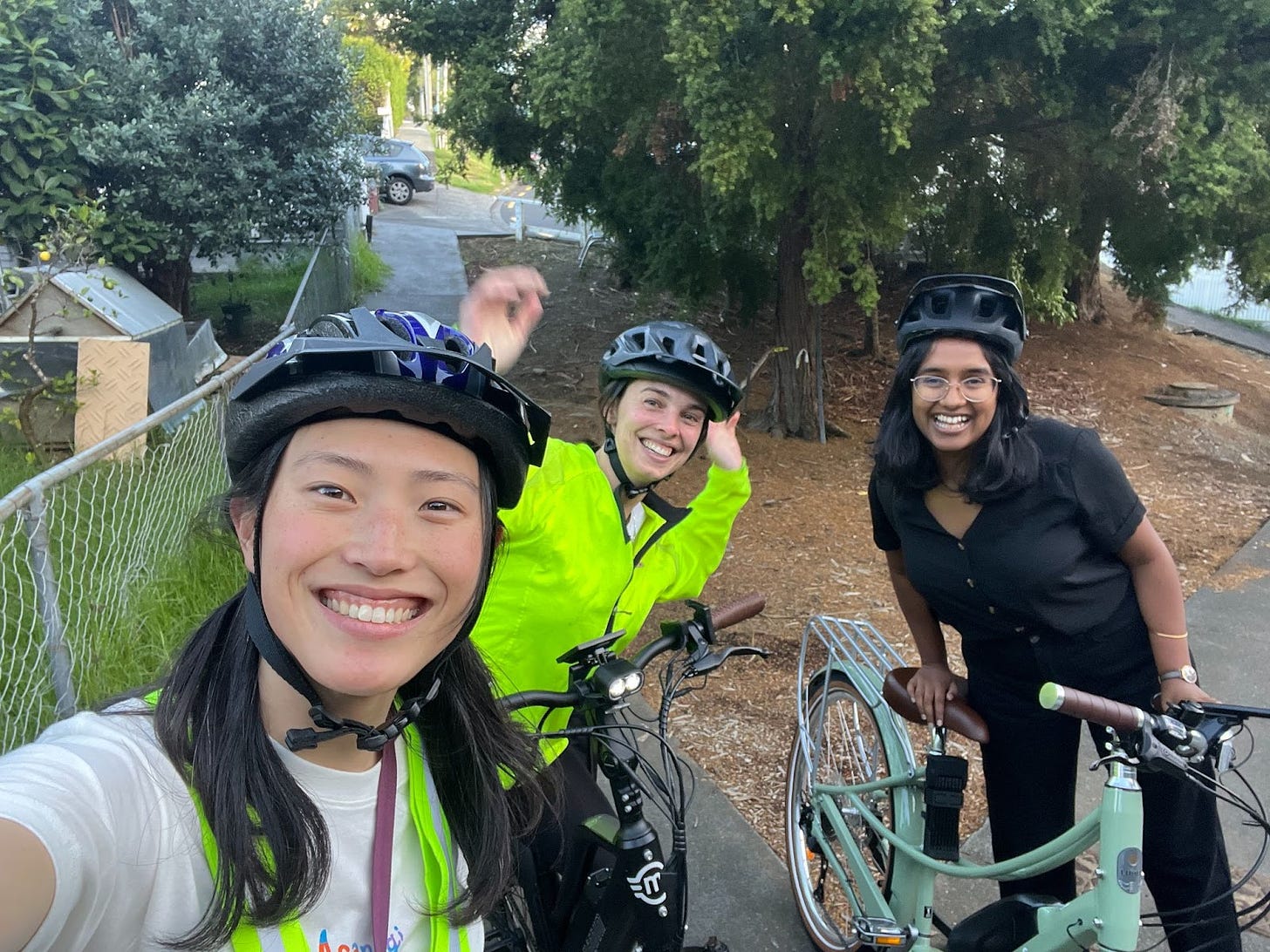 3 women smiling at the camera in bike helmets, with their bikes in front of them. Jenny has long black hair is holding the camera taking the selfie, Emily is in a high-vis jacket and raising her hands in excitement, and Dhanya is laughing while leaning on her sage green e-bike.