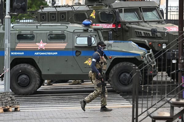 A soldier in camouflage, carrying a gun, walks past military vehicles on a city street.
