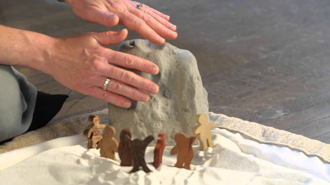 Wooden figures in sand gather around a model of Mt Sinai with two hands hovering around it