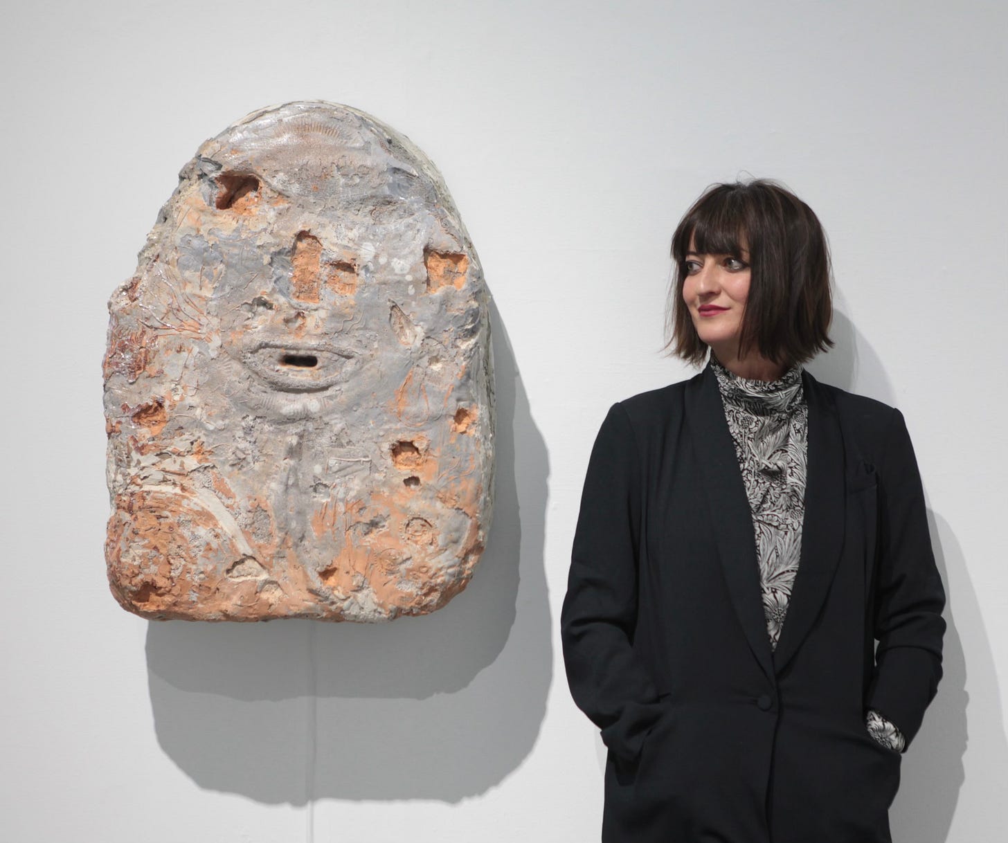 Mixed race woman with black hair in a black jacket stands beside a sculpture of rough earth tones hanging on a wall