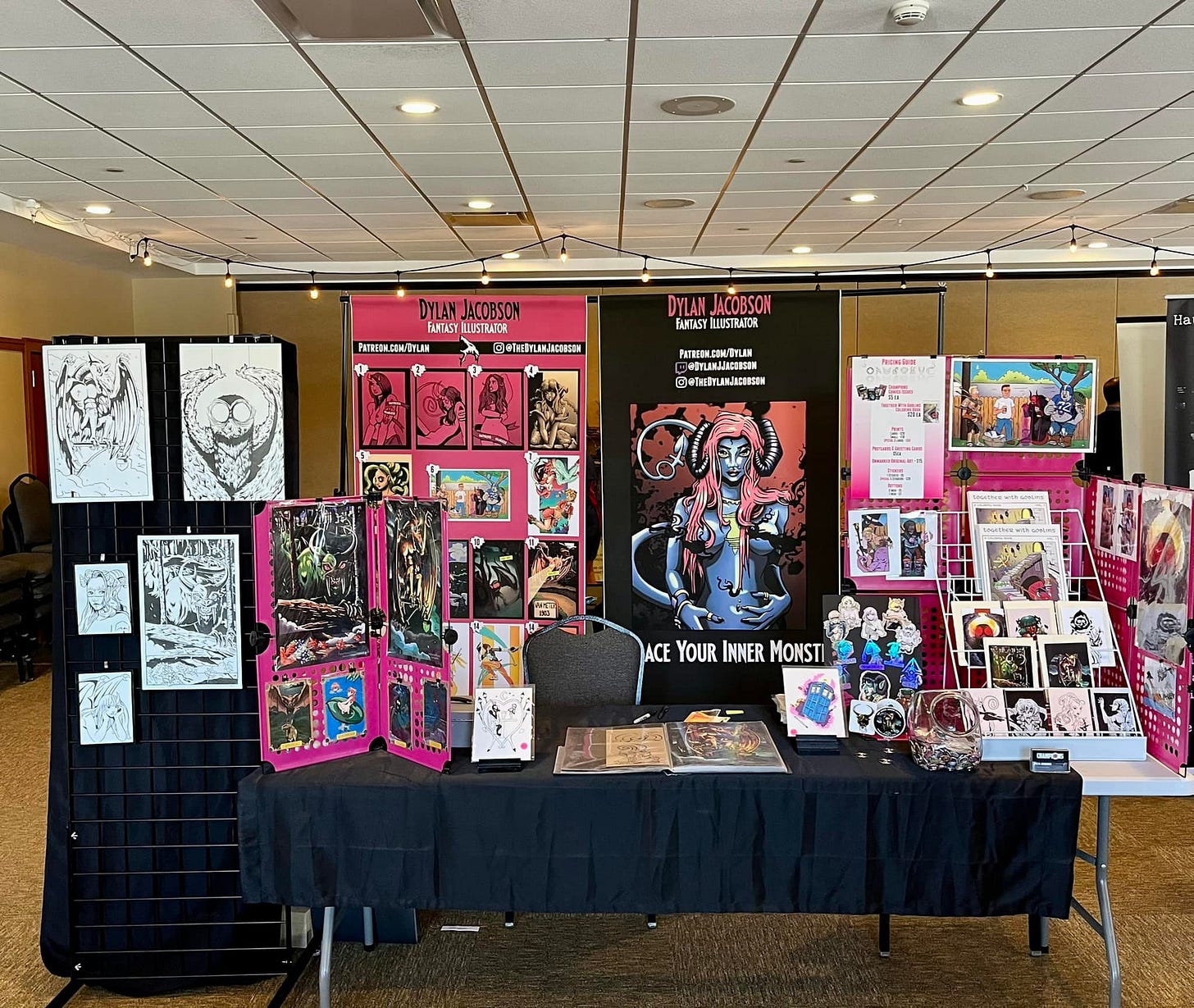 An elaborate convention set up featuring several pieces of original art, prints, and books.