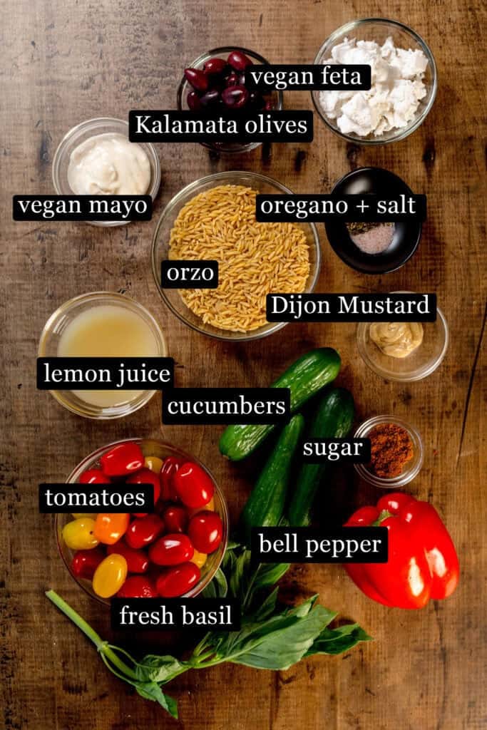 Ingredients for the greek orzo pasta salad in various bowls on a wood table. Black and white labels have been added to name each ingredient.