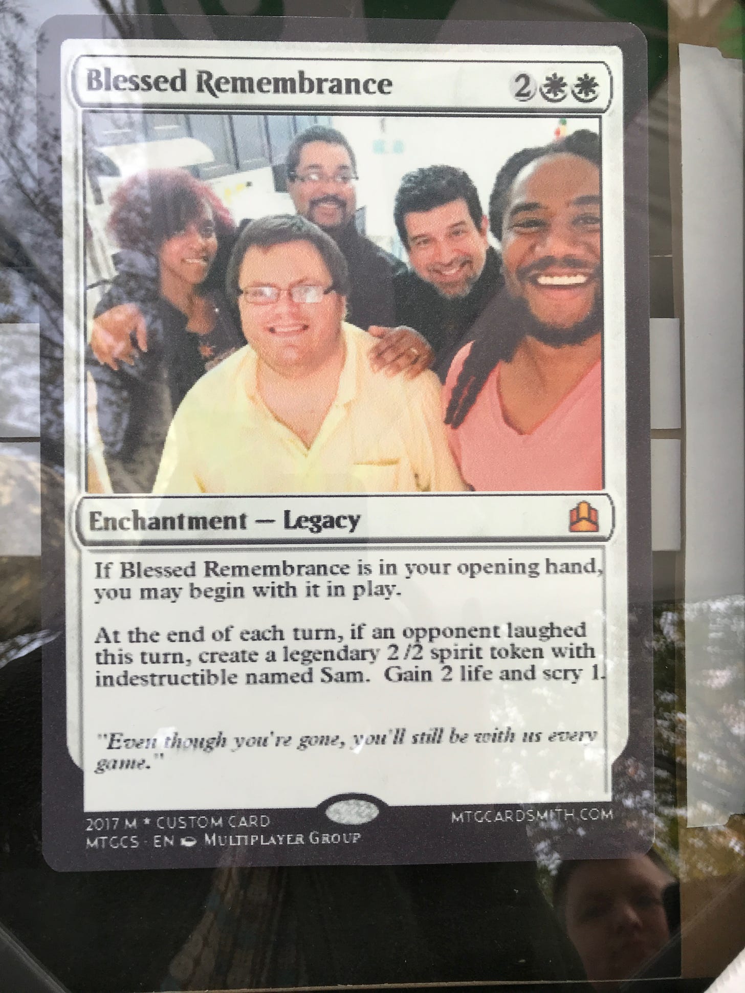 At the top of the card are the words "Blessed Remembrance." Under that is a photo of a bespectacled man in a yellow shirt (Sam) with four smiling friends. Below the photo are the words "Enchantment - Legacy." And then this text: "If Blessed Remembrance is in your opening hand, you may begin with it in play. At the end of each turn, if an opponent laughed this turn, create a legendary 2 /2 spirit token with indestructible named Sam. Gain 2 life and scry 1. 'Even though you're gone, you'll still be with us every game.'"
