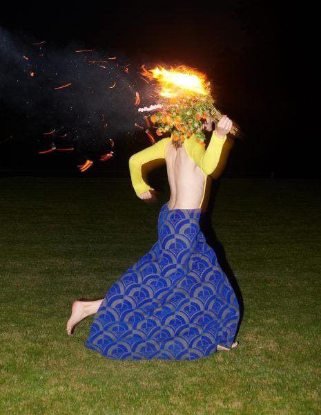 A femme wearing a floor-length dark blue skirt and a backless, yellow, long-sleeved shirt runs through the grass at night holding a flaming bouquet that blocks their face. 
