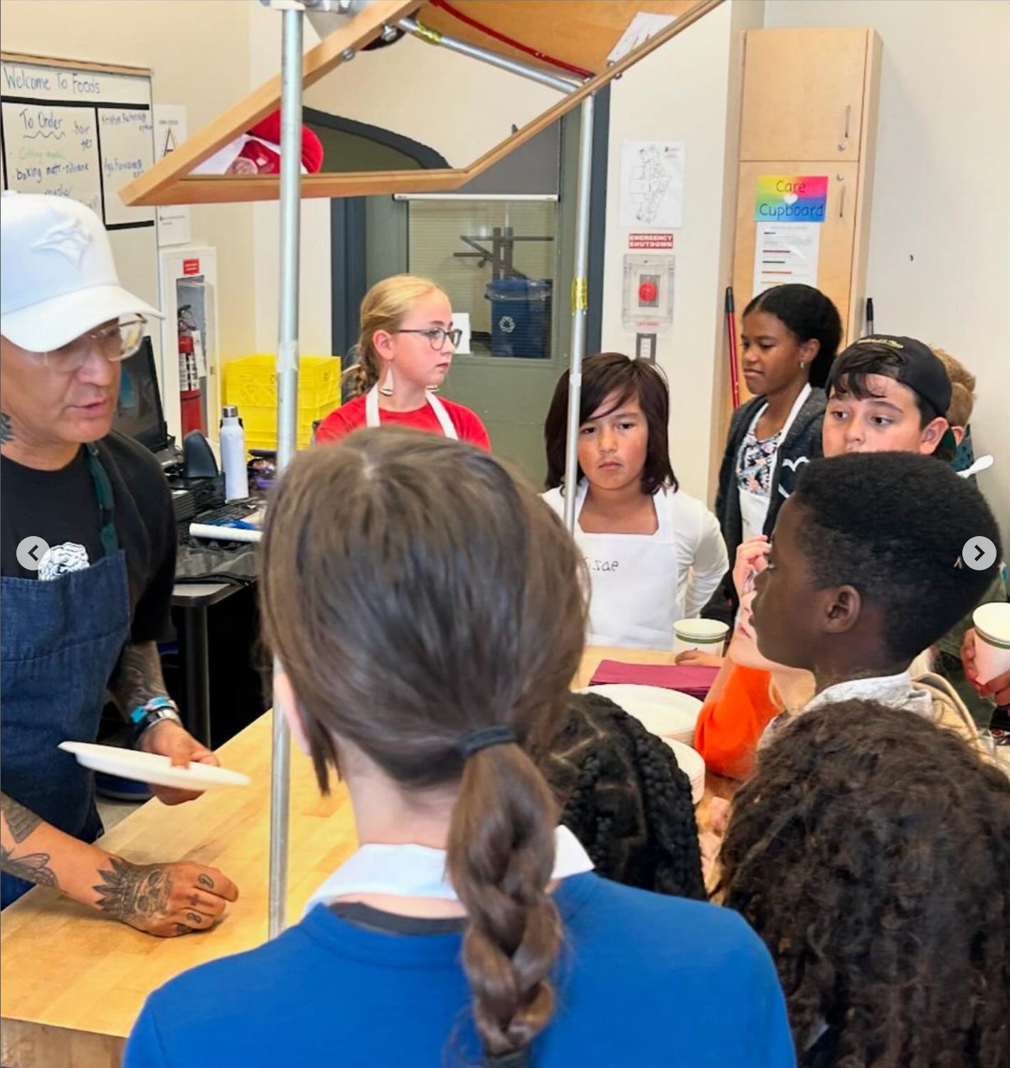 Chef Shane stands in a kitchen in front of 7 young kids of mixed races, teaching them about indigenous cooking.