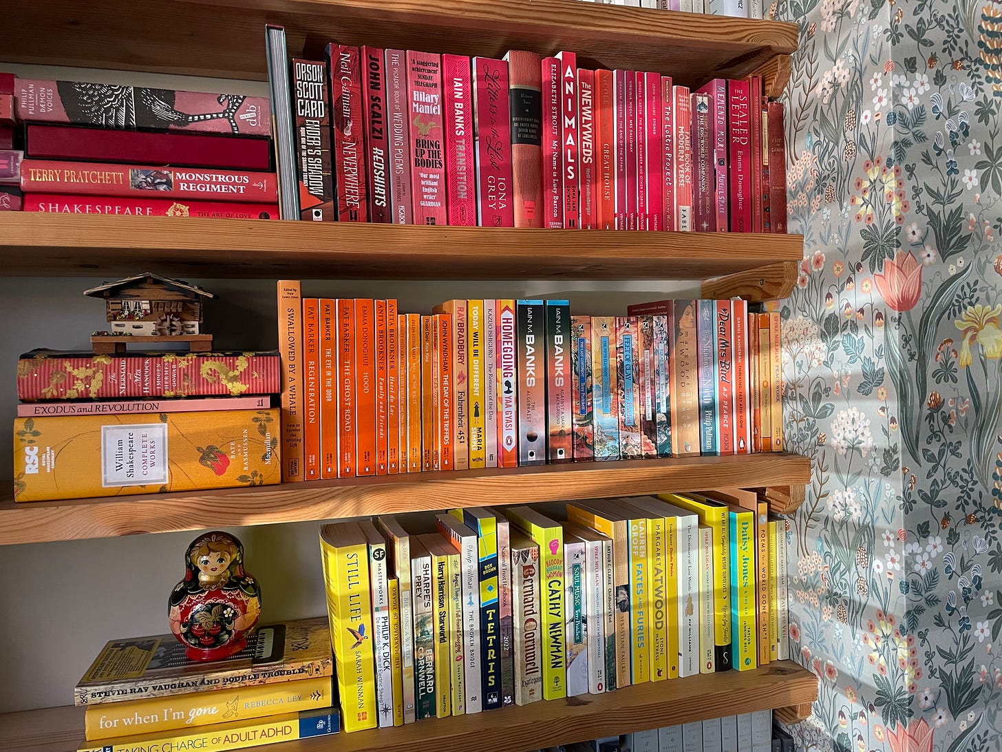 A photo of my bookshelves. Three pine shelves are visible, one with red books, one with orange and one with yellow. You can also see floral wallpaper on the wall, and the light shining in through the slats of the blinds. 