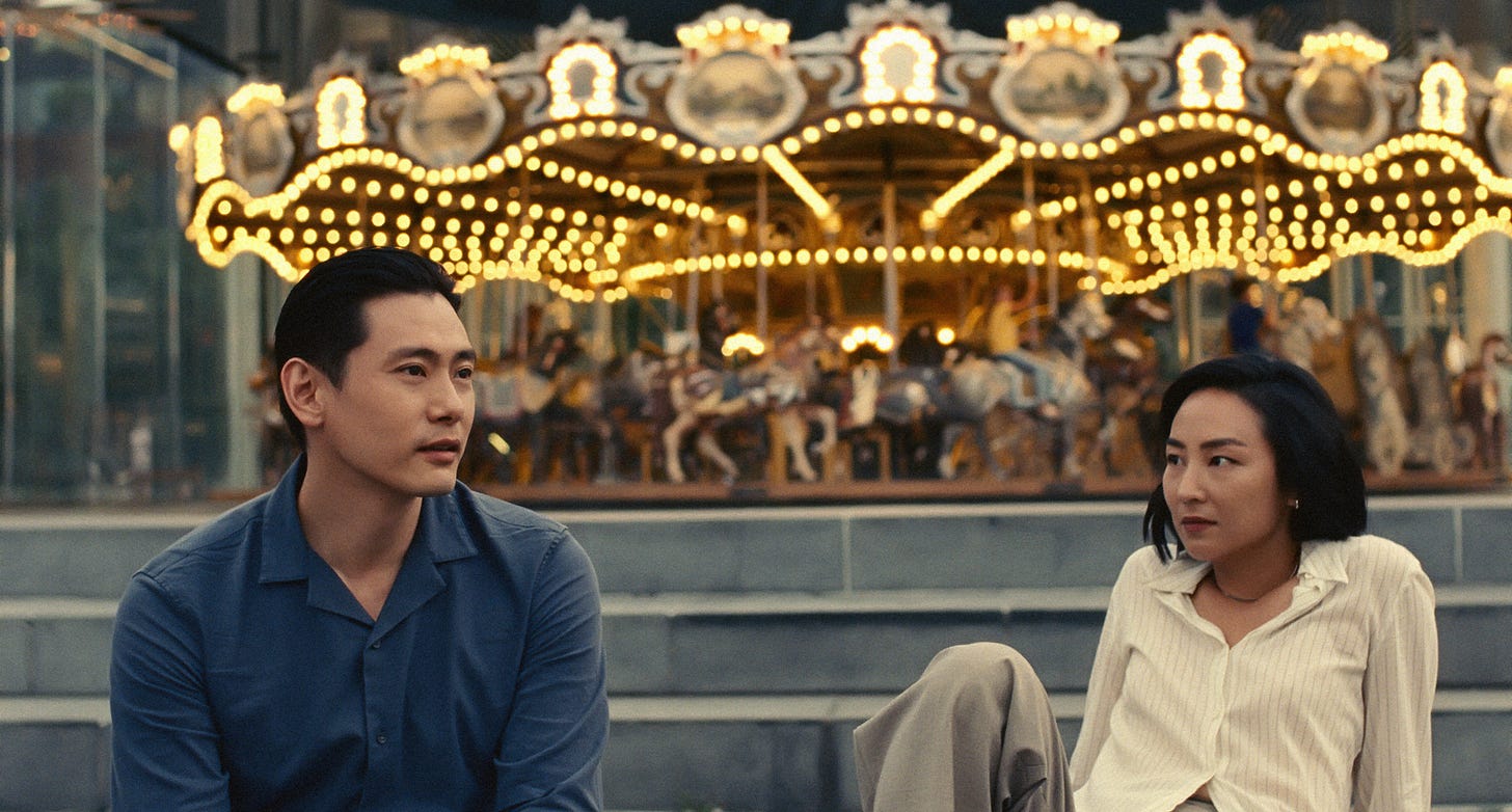 Photo extraite du film Past Lives, où l'on voit Nora à droite et Hae Sung à gauche, avec en fond un carousel illuminé. Elle le regarde, il regarde au loin.