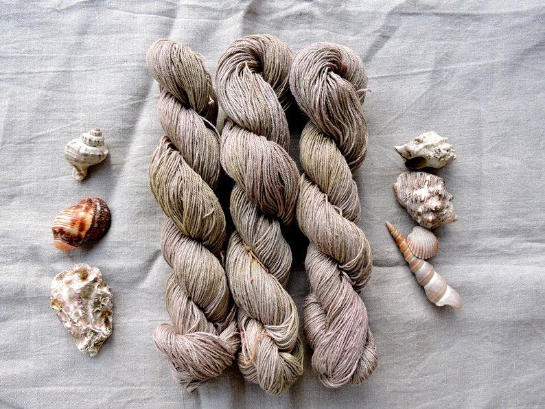 May include: Three skeins of yarn in a light brown and gray color. The yarn is arranged in a vertical stack. The yarn is surrounded by seashells in various colors and shapes.