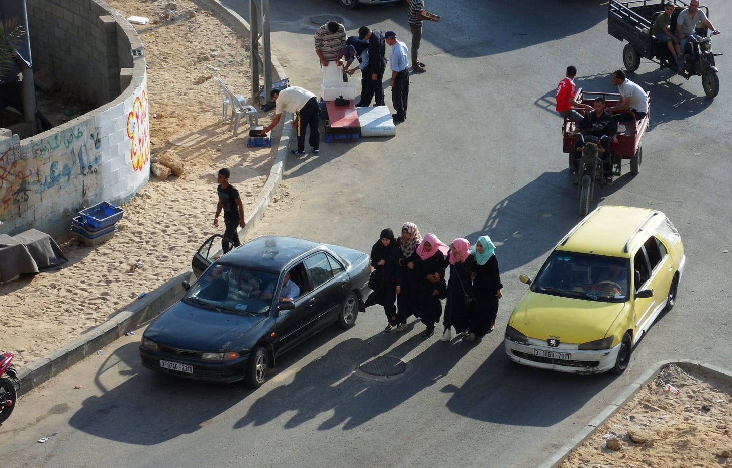 A group of women in headscarves walk among cars and motorcycles on a busy afternoon street