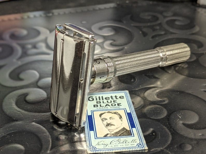 1962 Gillette Slim Adjustable Vintage Safety Razor with packet of tradmarked disposable Gillette Blue Blades. The razor is metal, probably stainless steel, with a shiny silver finish. The blade packet is made of two-toned light and dark blue paper and bears the image and signature of King Camp Gillette.
