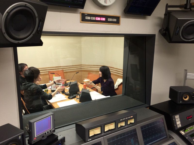 View of a recording studio of radio broadcaster Nippon Cultural Broadcasting. In the foreground are recording studio control panels. Beyond the window is a recording room with three people seated around a table that has microphones and papers on top of it.