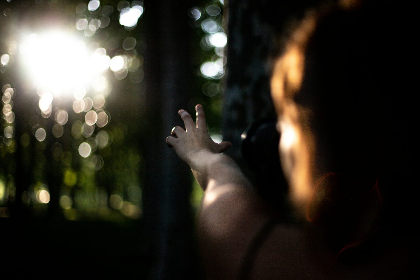Ripresa sfocata di una figura umana femminile che protende il braccio sinistro in avanti, in mezzo a un bosco, puntando verso una fonte luminosa che sta sullo sfondo in alto a sinistra.