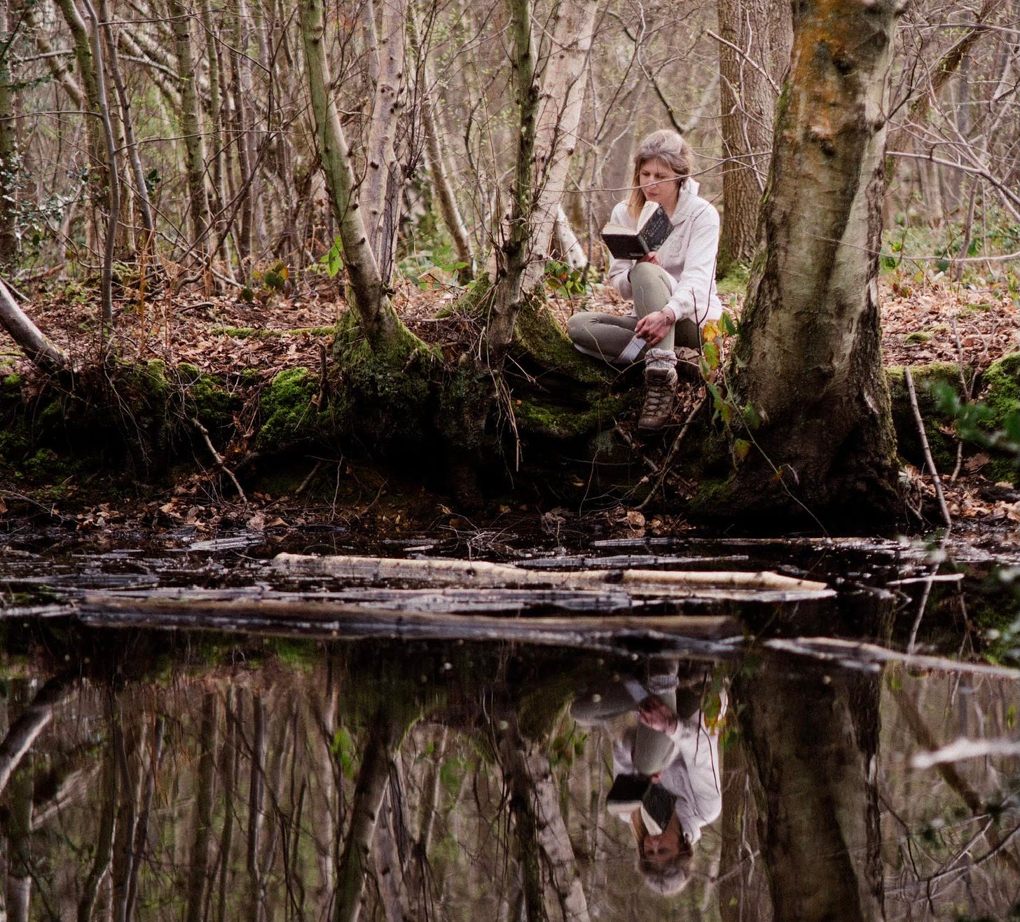 Middle-aged white woman seated in a spring forest at water's edge, reading a book, her image reflected in the still surface.