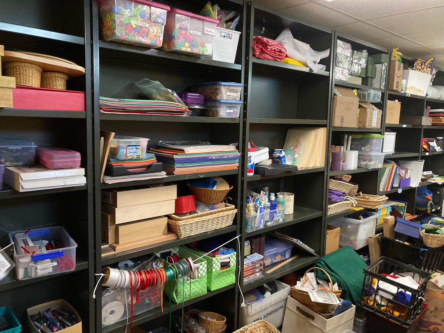 A photograph of a supple closet packed with papers, bins, and baskets on black metal shelves. It is overflowing with items on the floor, as well.