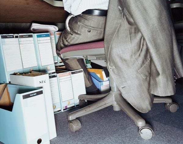 This may contain: a man sitting in an office chair next to a desk with several files and folders on it