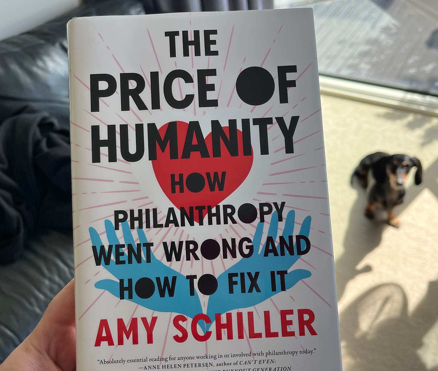 In the foreground a hand holds a book titled The Price of Humanity, how philanthropy when wrong and how to fix it, by Amy Schiller. In the background a blurry dachshund looks on.