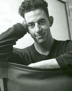 Black and white image of Jonathan Larson sitting on a chair backwards, smiling. We can only see the back of the chair, part of his chest, shoulders, and face. He seems to be wearing a black shirt. One arm is resting fully on the back of the chair, and with the other arm, he props his elbow onto the back of the chair and rests is hand on on the side of his face, so that he can lean on his arm. The hand on his face is covering his ear. 