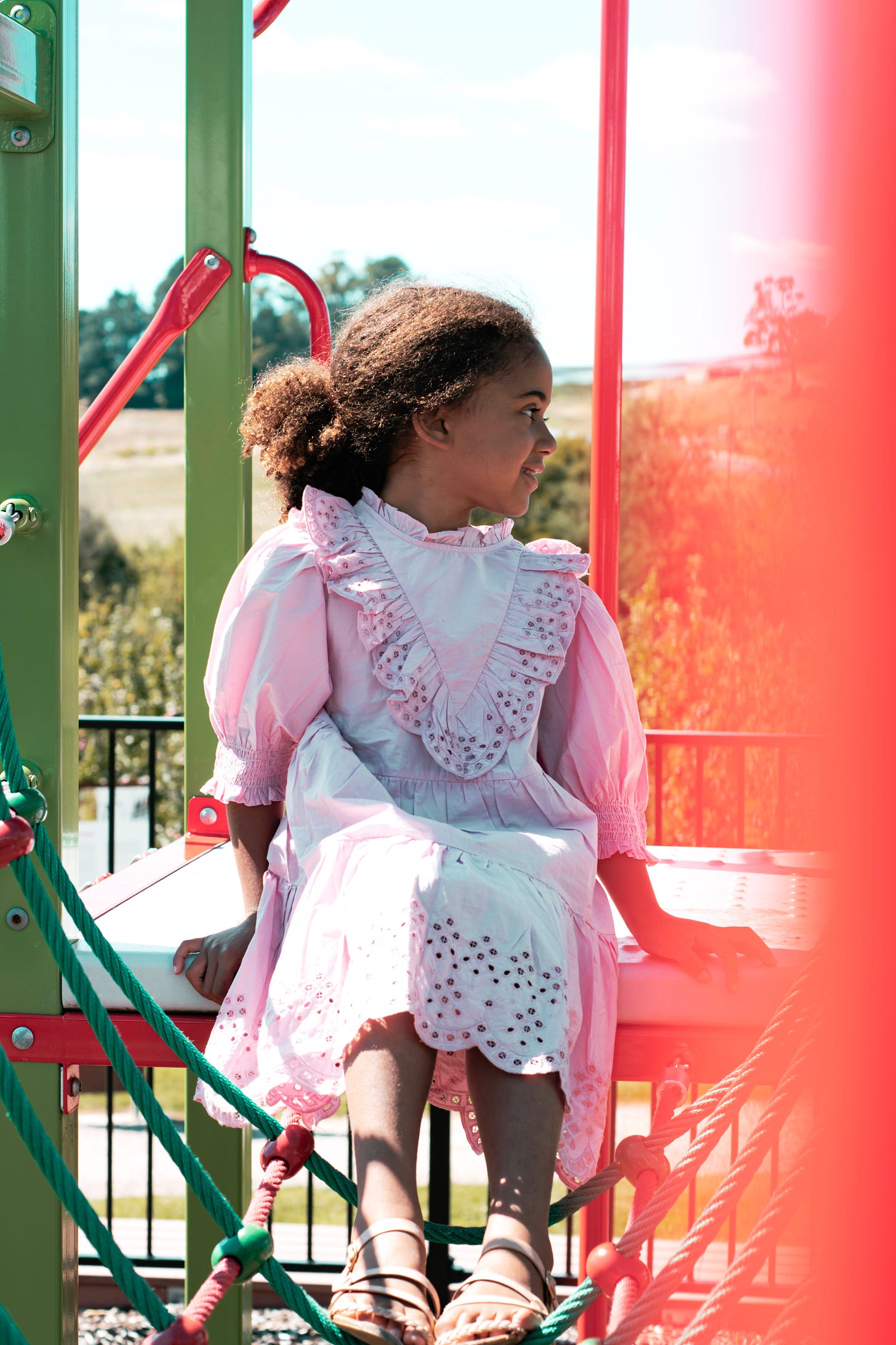 Young Black girl sits on red playground gym platform. She is smiling, head to side.