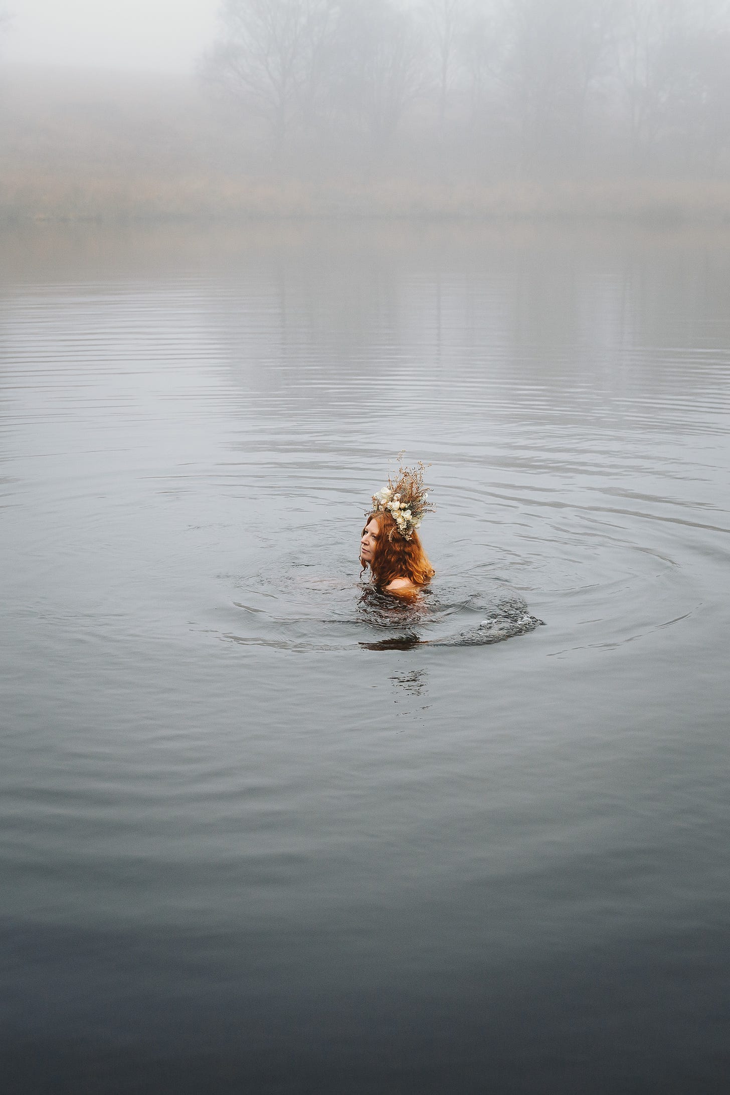 A woman wears a flower crown and is up to her neck in water. The pond is misty and there are faint trees in the background.