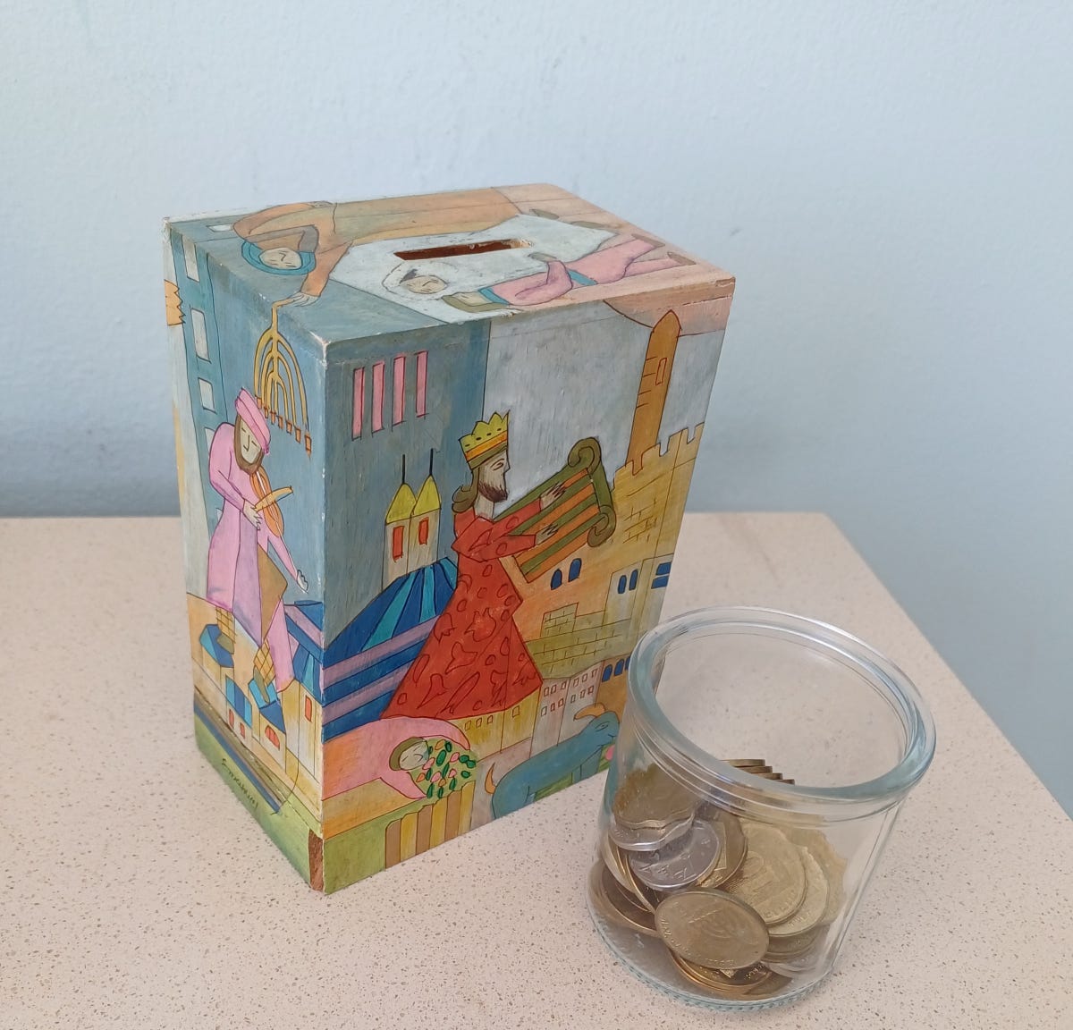 Hand-painted pushke on table with little glass jar of coins beside it