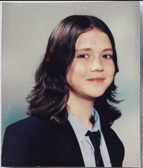 Young Janelle aged approx 13 in her official school head shot. She has a hint of a smile, is looking into the camera. Wearing a navy blazer and tie. She has brown eyes and shoulder length brown hair with flicky layers