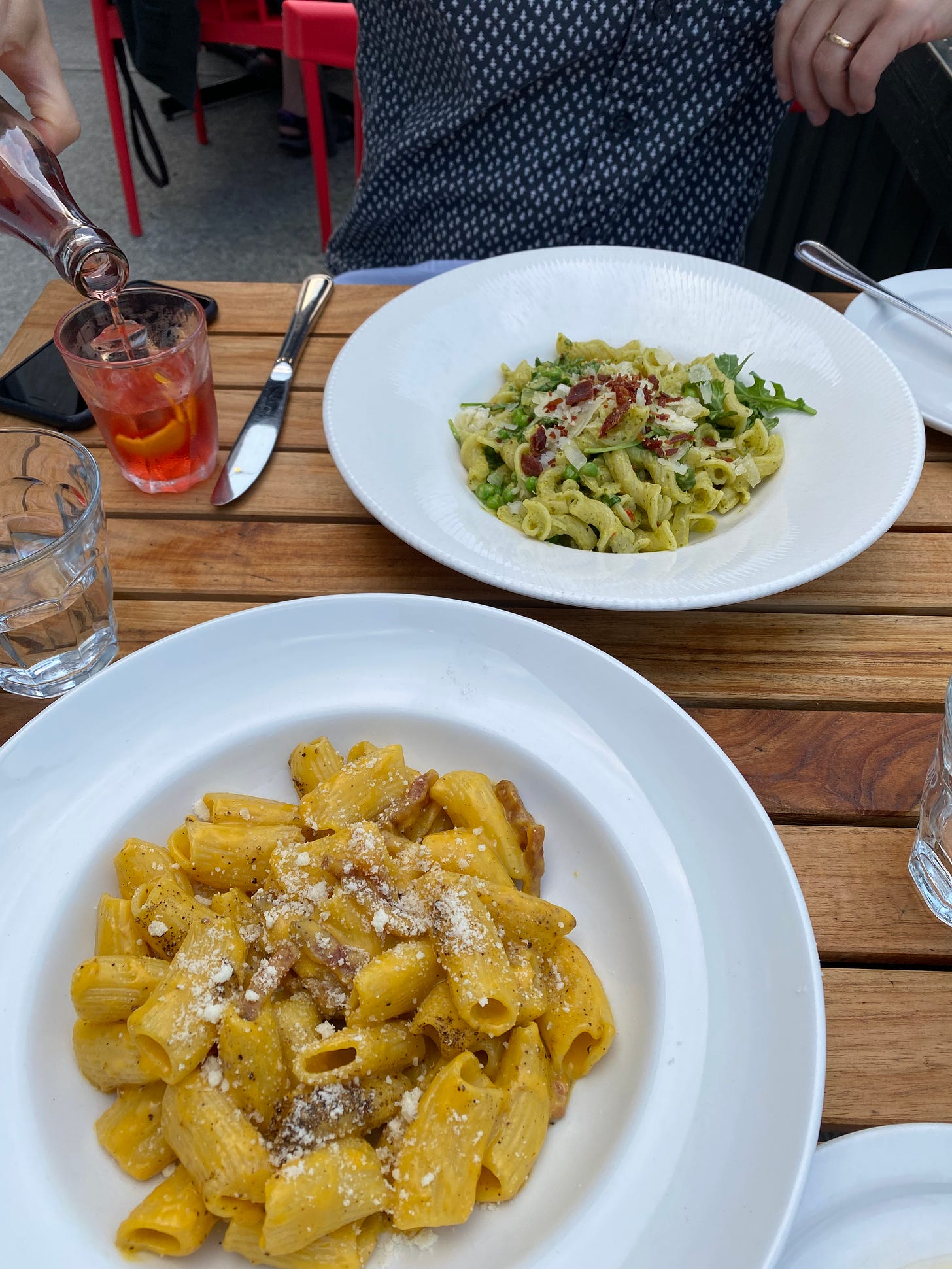 Two white bowls of pasta on a patio table. In the foreground is rigatoni carbonara, with crumbles of parmesan on top. On the other side of the table is campanelle in a green sauce with prosciutto, arugula, peas, and parmesan. Jeff's hand is in the left of the frame, pouring a red bottled cocktail into a small glass with an orange rind.