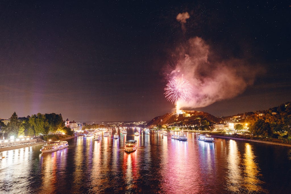 Lots of boats light up in different colours sailing towards the camera on a wide river, with fireworks going off on a hill in the background next to a fortress