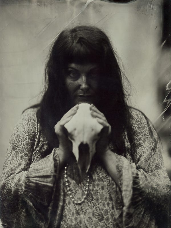 An old, shadowy image of a dark haired woman in a flowing floral dress holding a sheep's skull under her chin. Her face is in shadow and she wears an intense expression. The surface of the photo is distressed