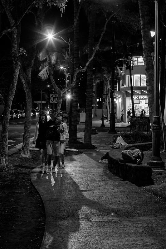 Monochrome night time photo taken on Waikiki's Kuhio Avenue. Tourists backlit by streetlights and a passing automobile walk toward the camera midframe, casting shadows intermingled with those of the palm trees at the left of the frame. A homeless man sleeps on a low, curved, concrete wall to the right of the frame, next to a lamp post and another palm tree. In the background is one of Waikiki's ubiquitous brightly-lit ABC stores with several people silhouetted in the entrance.