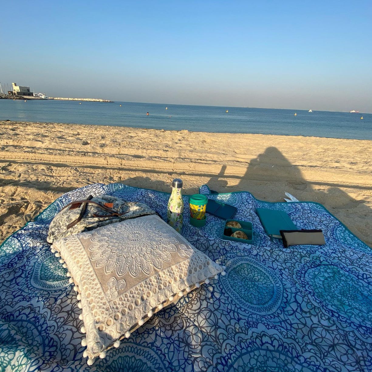Photo of the picnic at the beach the writer went to. There is a floral blue and white picnic blanket topped with a floral cream and white pillow, a colorful thermos bottle and coffee cup, an open container with breakfast in it, a journal, pencil case, and a silver floral backpack. Photo of the picnic at the beach the writer went to. There is a floral blue and white picnic blanket topped with a floral cream and white pillow, a colorful thermos bottle and coffee cup, an open container with breakfast in it, a journal, pencil case, and a silver floral backpack.