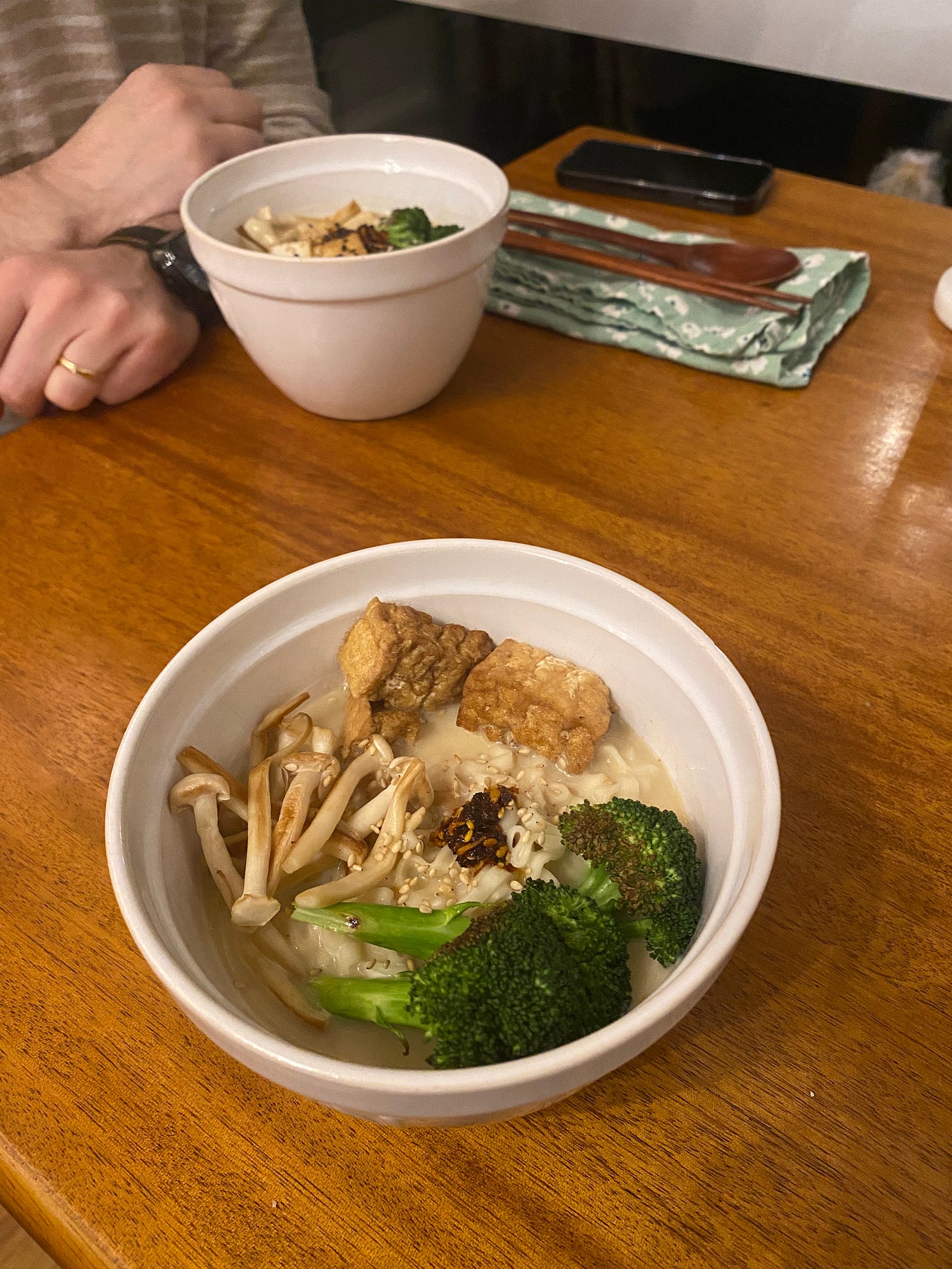 Two white bowls of the ramen described above: noodles in a creamy broth topped with pan-fried mushrooms and broccoli, pieces of puffed tofu, and chili crisp. Jeff's hands rest on the table behind his bowl.