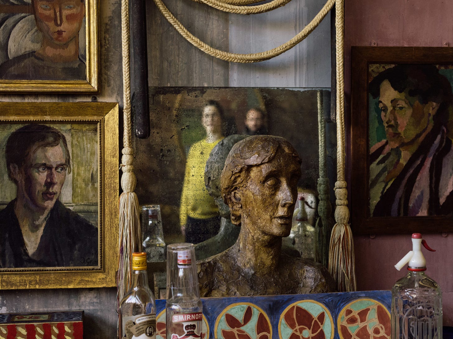 Crowded scene with multiple paintings in the background, a bust of Virginia Woolf in the center, old bottles and decorative items scattered about. 