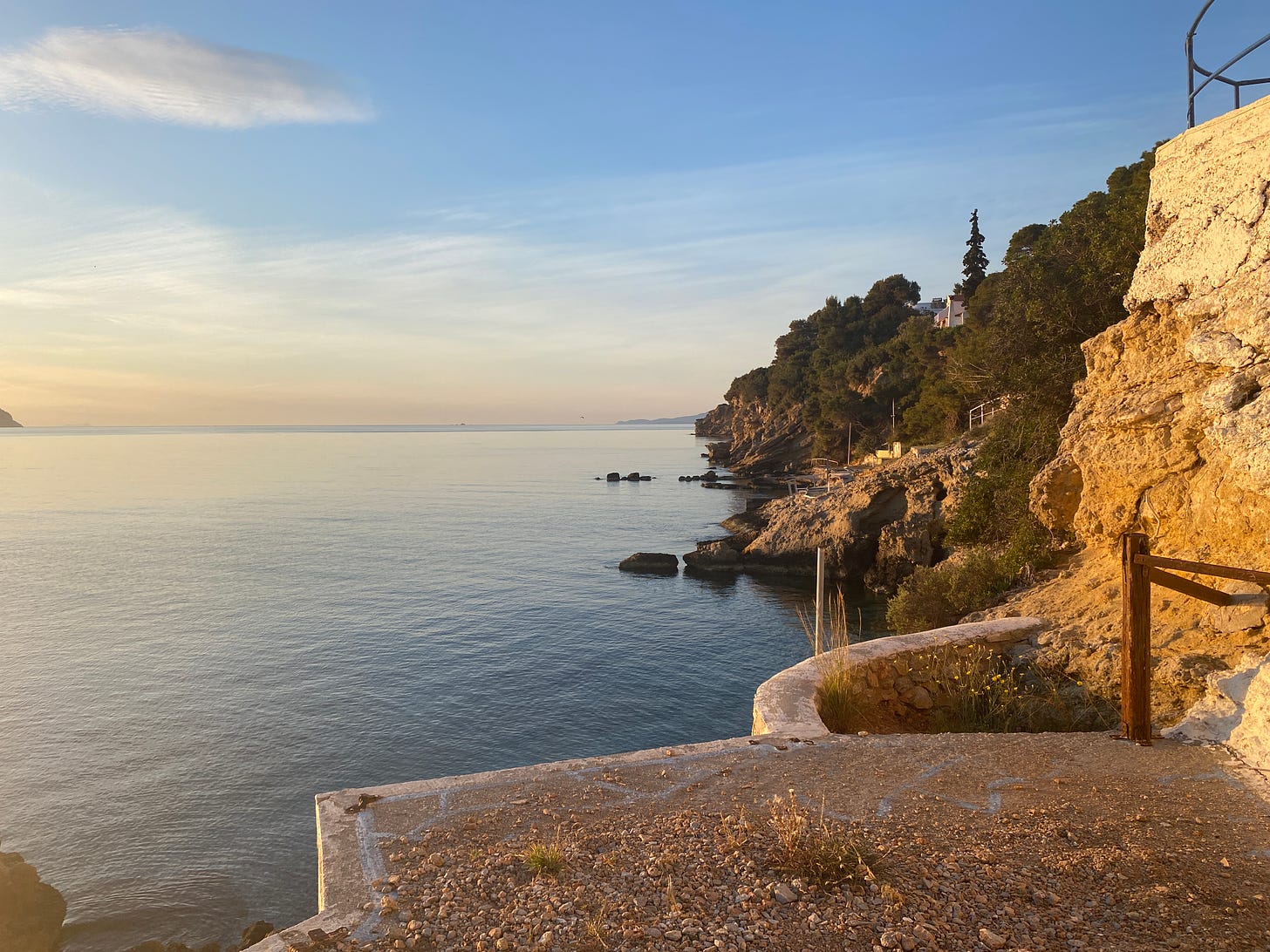 Yellow light hits the rock face with pastel coloured water and a pale pink sky close to the horizon and a bluer white fluffy cloud sky towards the top of the frame.