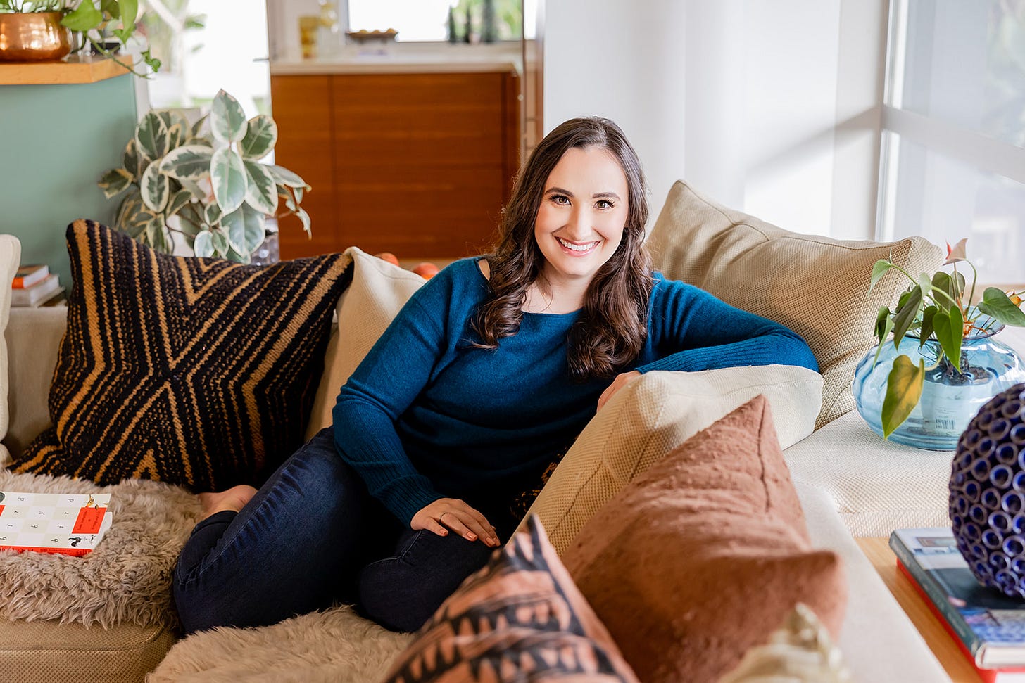 Taylor is a white woman with long dark brown hair. She is lounging on a couch and smiling at the camera.