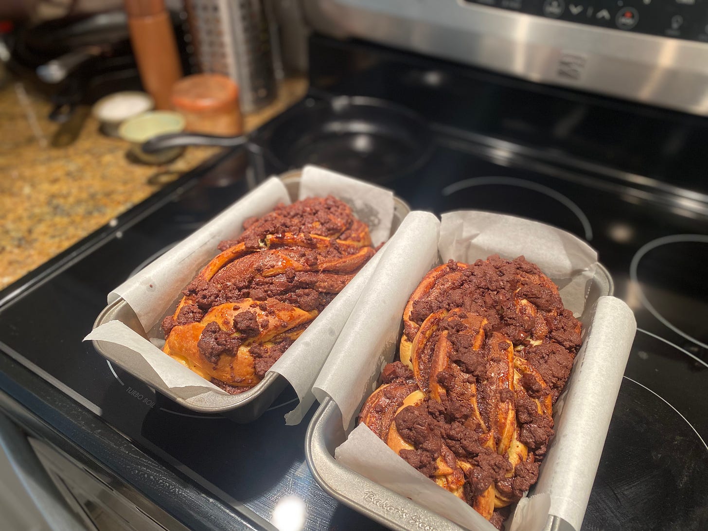 The two pans of babka, fresh out of the oven and resting on the stovetop. They are risen and browned from the oven, and there is a chocolate streusel topping which has puffed up as well.