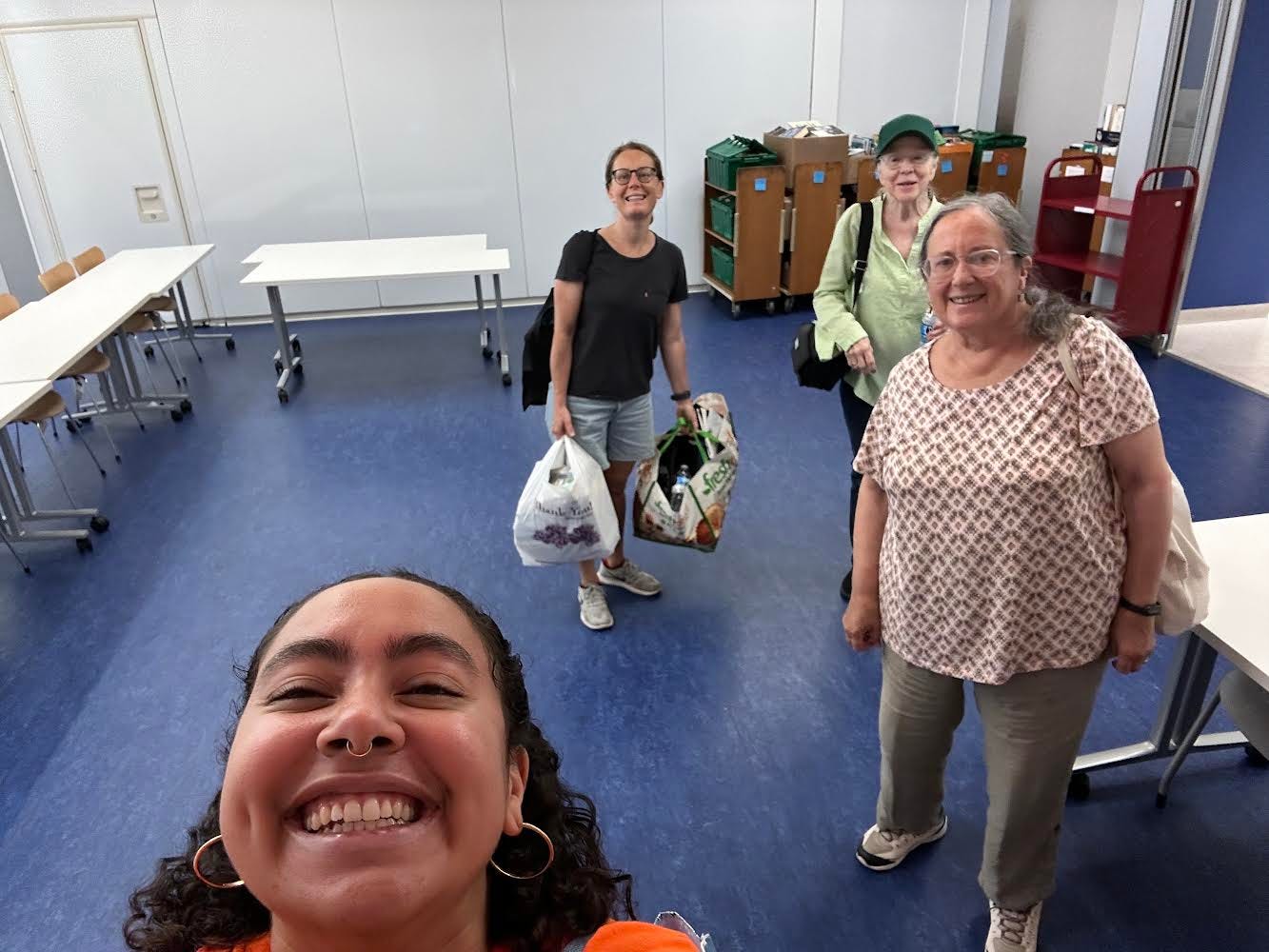 Selfie-style photo with Keren (who is clearing the one holding the camera) foregrounded, and three other friends of the library in the background. They are in a room with empty tables around the edges and appear to be packing out.