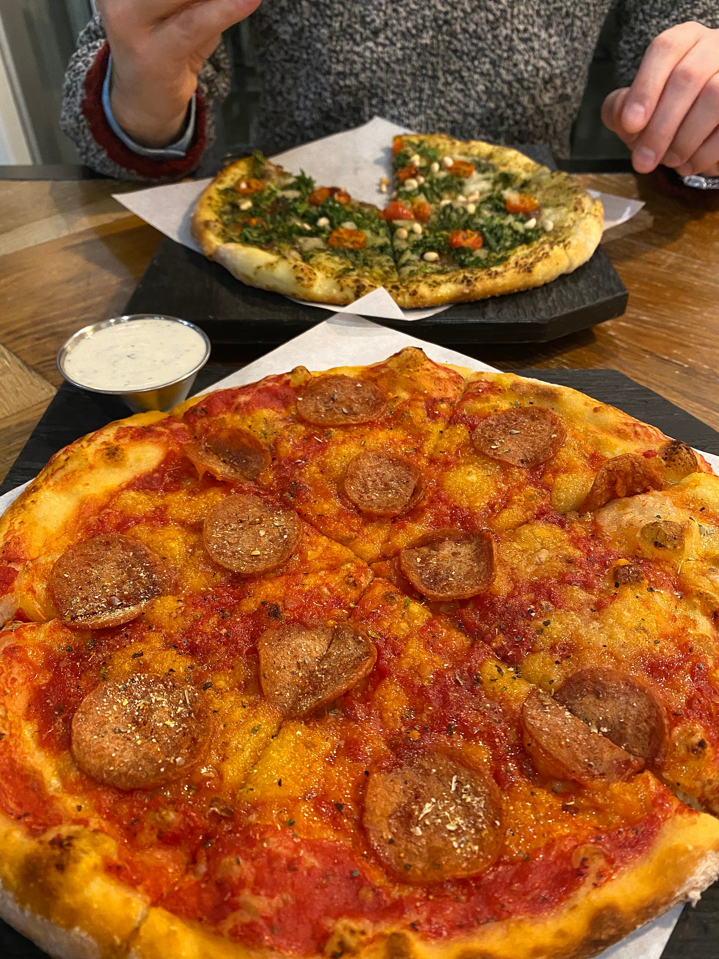 Two pizzas on black serving platters. In the foreground is the pepperoni, dusted with seasoning and with a small ramekin of ranch. In the background, Jeff is holding a slice of the ultraviolet, a pesto based pizza with lots of kale, and oven-dried grape tomatoes.