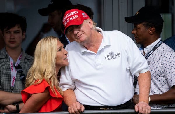 Former President Donald J. Trump, wearing a white polo shirt and a red MAGA hat, leans over toward Representative Marjorie Taylor Greene, who is talking near his ear.