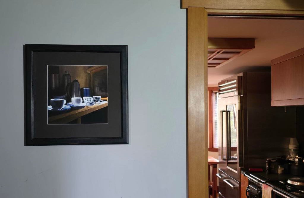 Framed art print of four espresso cups illuminated by a shaft of sunlight. The print hangs in a hallway outside an opening where a sunlit kitchen can be seen.