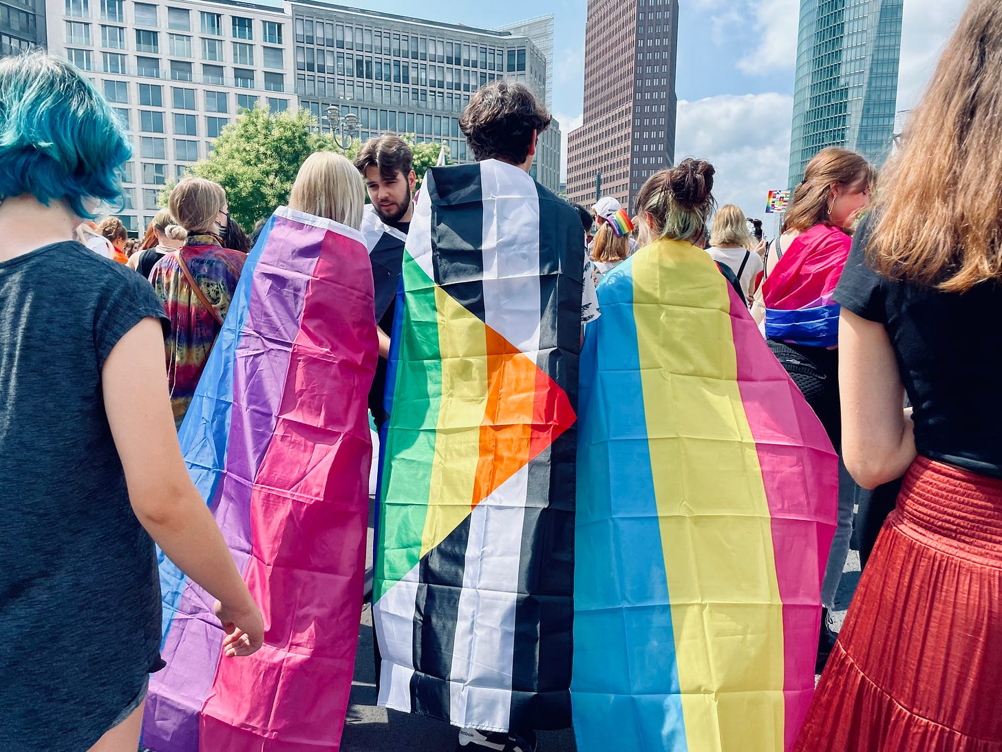 A group of people wrapped in Pride flags, including the bisexual, nonbinary, and pansexual flags, stand together in a lively crowd at Berlin CSD 2023. Tall buildings and trees are visible in the background.