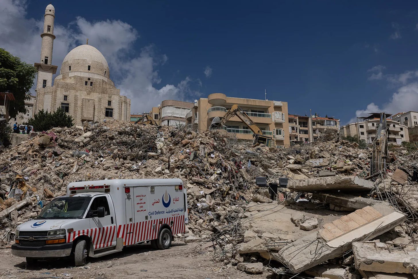 An emergency vehicle is seen amid a scene of building rubble and destruction. An excavator can be seen on top of the rubble. Several intact buildings and the dome of a mosque are seen farther in the background.