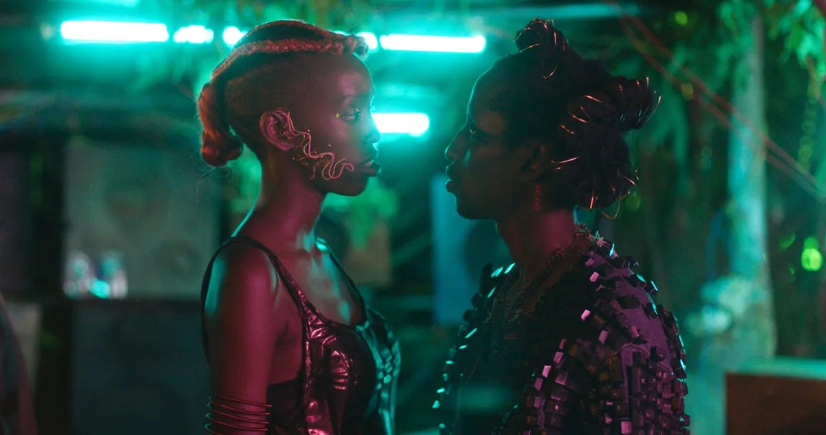 Film still from Neptune Frost showing two Black people bathed in pink and purple light facing each other against a futuristic backdrop of emerald green lights.