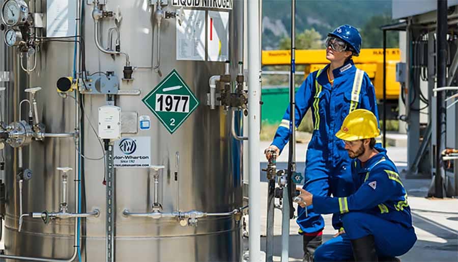 Two workers in blue uniforms and safety gear diligently operate equipment at an industrial site focused on sustainable practices, near a large container labeled "Liquid Nitrogen," showcasing their contribution to green jobs.