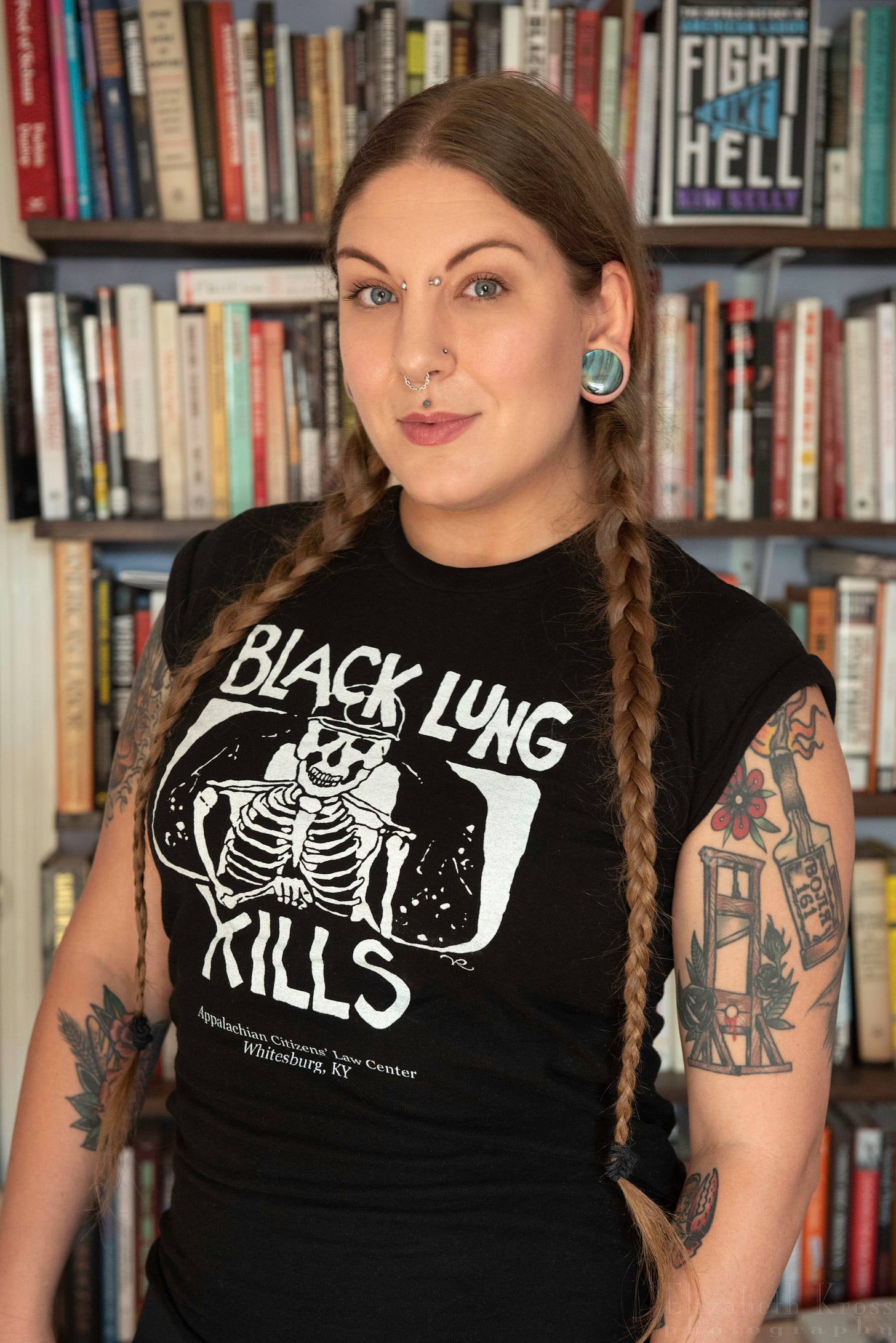 A portrait of Kim Kelly, an author, photographed from the waist up standing in front of a bookshelf with her book, Fight Like Hell, featured prominently but slightly out of focus over her left shoulder. Kim is a white woman with reddish brown hair styled into two French braids. She has numerous facial piercings and tattoos on her arms, including one of a molotov cocktail and another of a guillotine. She is wearing a sleeveless black t-shirt that says “Black Lung Kills” across the chest.