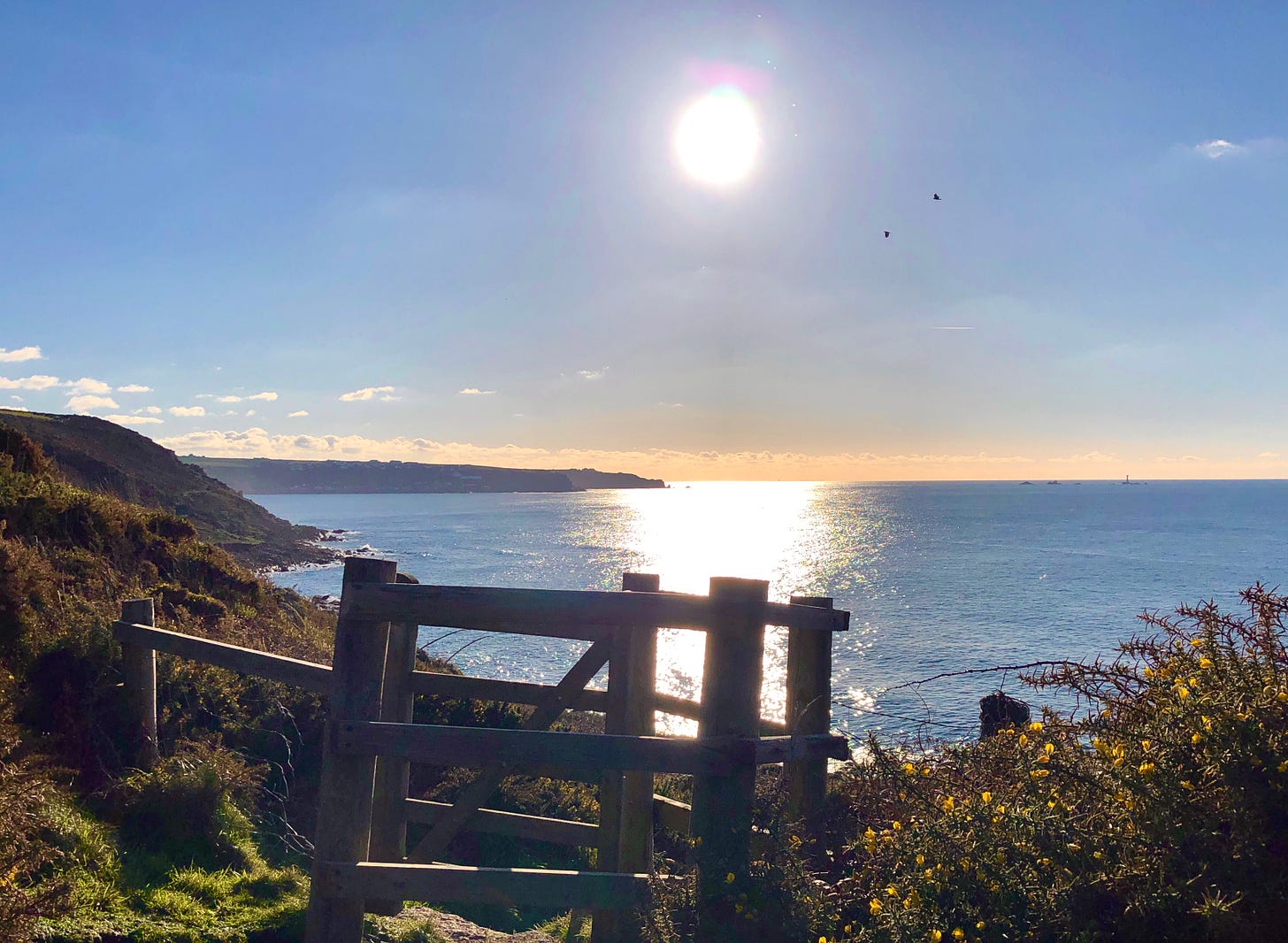 A wooden gate on the coast path. The sun is shining brightly and reflecting off the blue sea.