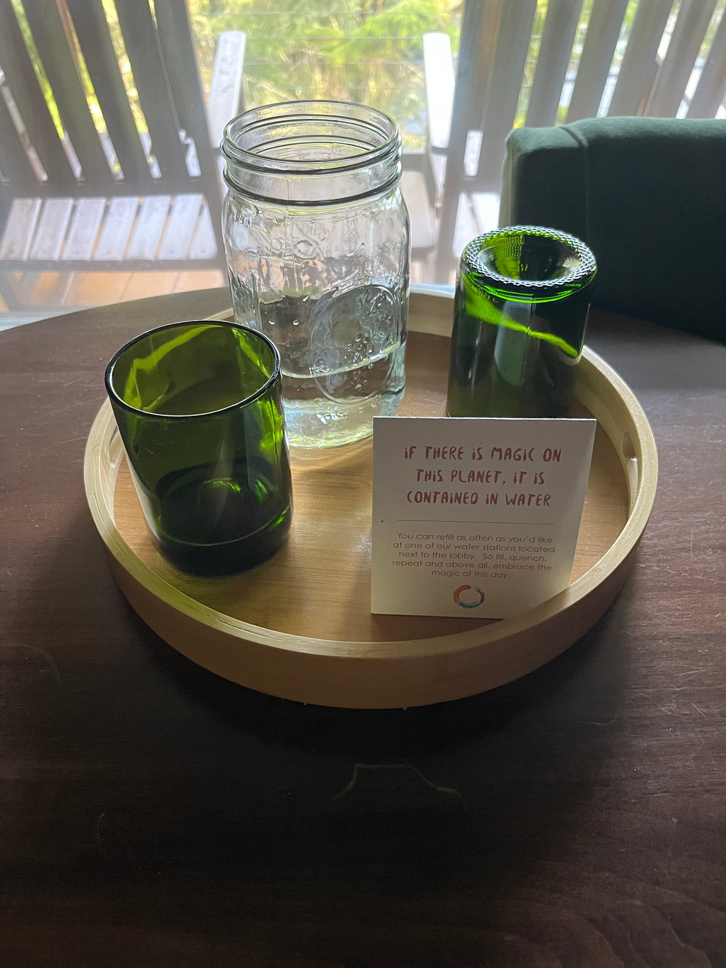 A bamboo tray with two green glasses and one glass mason jar of water sits on a table in a hotel room. 