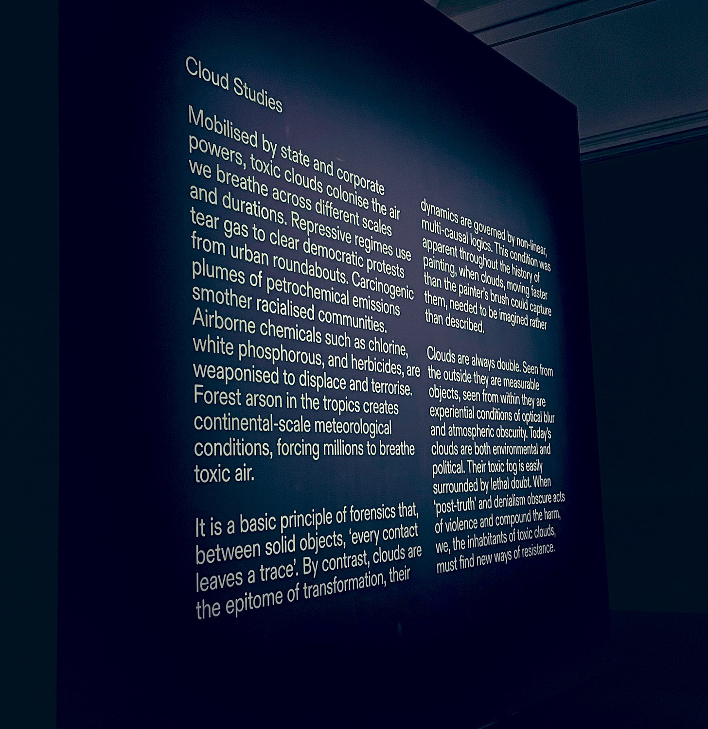 the exhibit information on the wall on “cloud studies” . The information is in white text in a dark room.