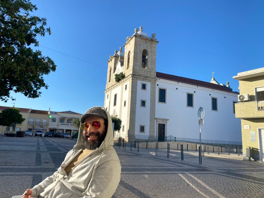 Man with bear, shades and a hoody sat in a square with a church tower behind