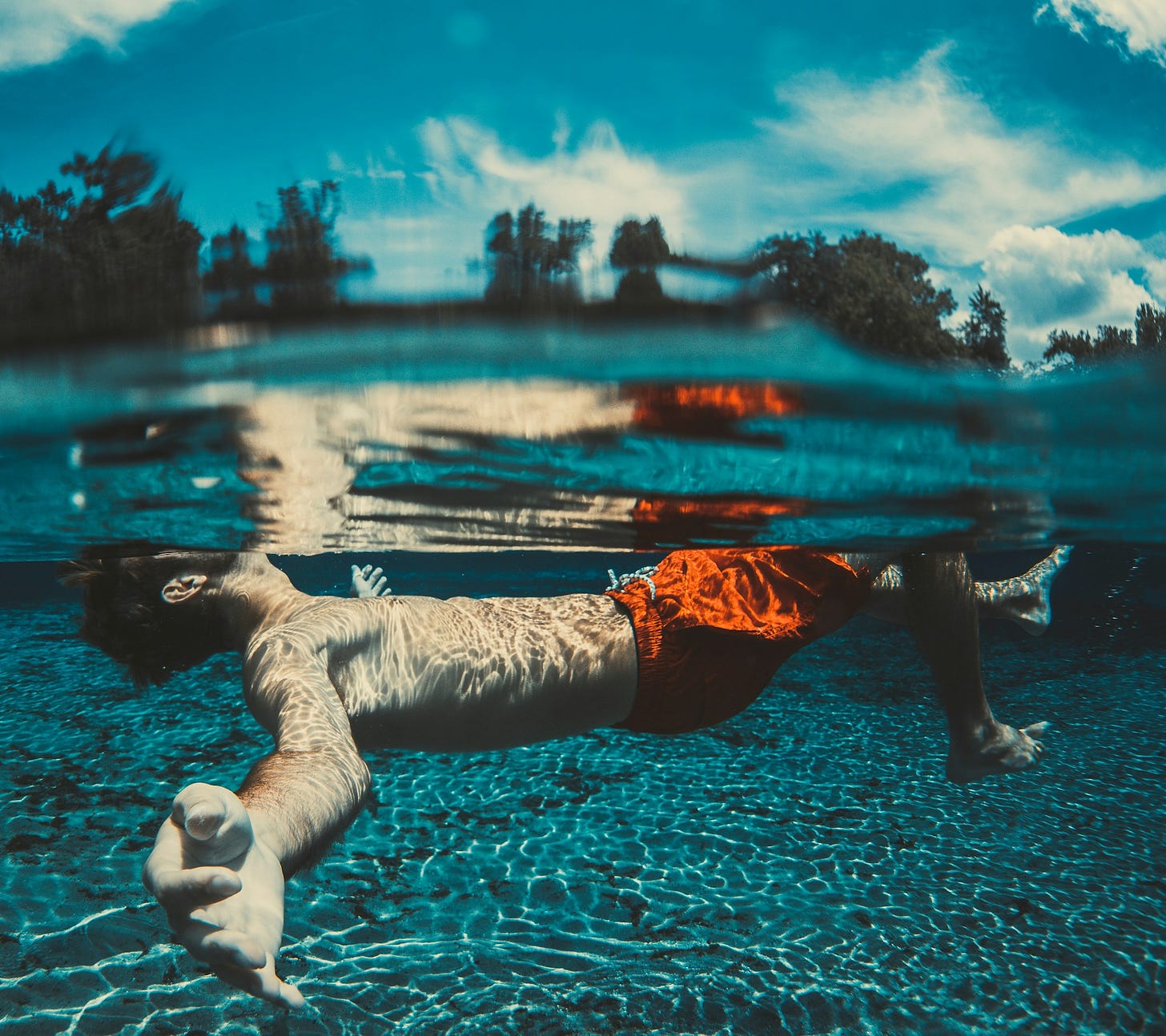 A young man wearing orange swim shorts floats on his back in the water, with trees and blue sky in the background.